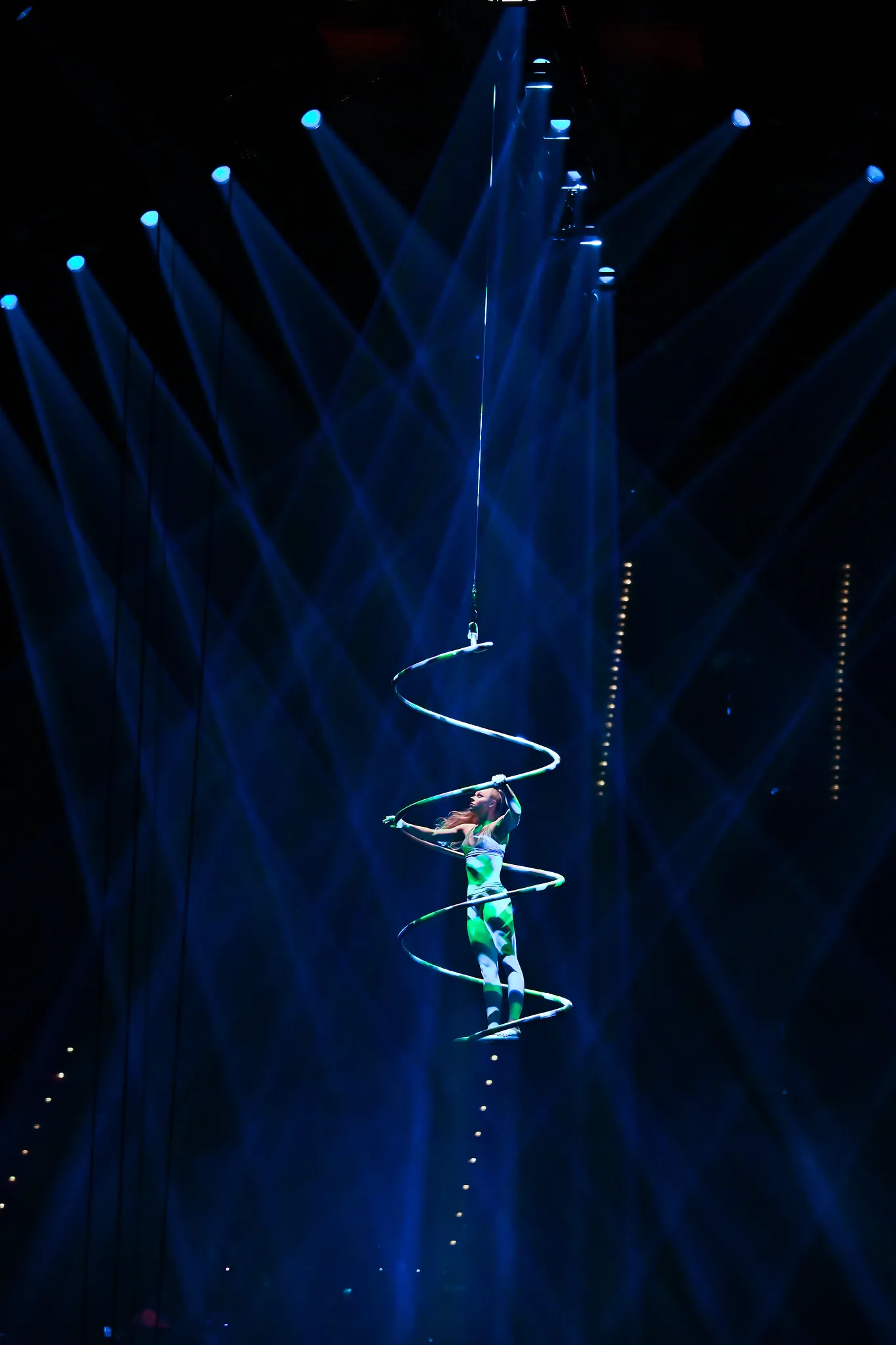 Aerial-Artistin in spiralförmigen Luftringen, schwebend im blauen Bühnenlicht, Kulturfotografie einer Zirkusperformance.