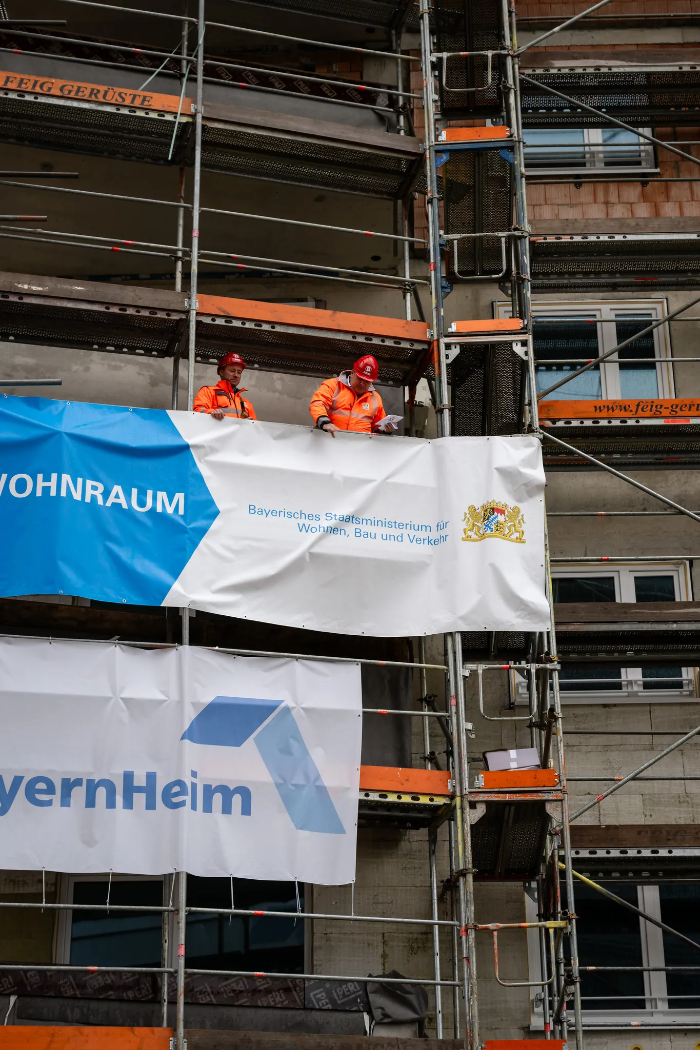 Zwei Bauarbeiter auf Gerüst vor BayernHeim-Wohnraum-Banner