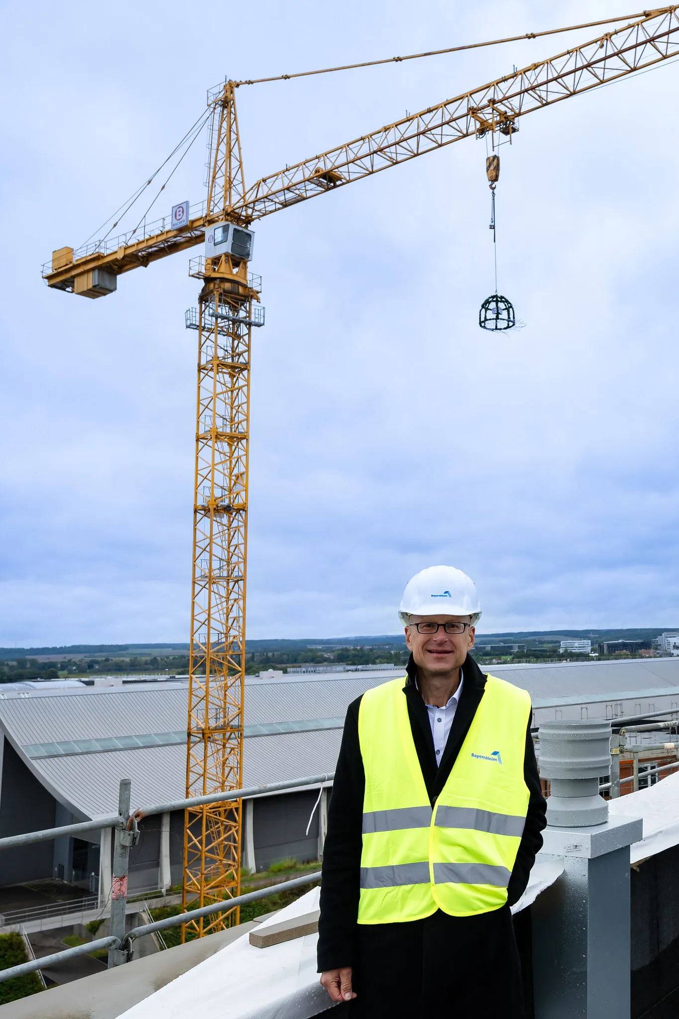 Projektleiter mit BayernHeim-Bauhelm vor gelbem Kran auf dem Dach