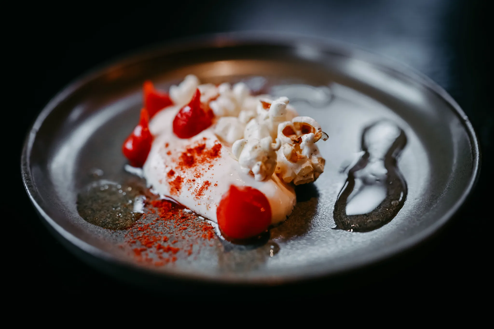 Fine-Dining-Dessert mit Popcorn, Tomatengel und Sahnecreme auf dunklem Teller.