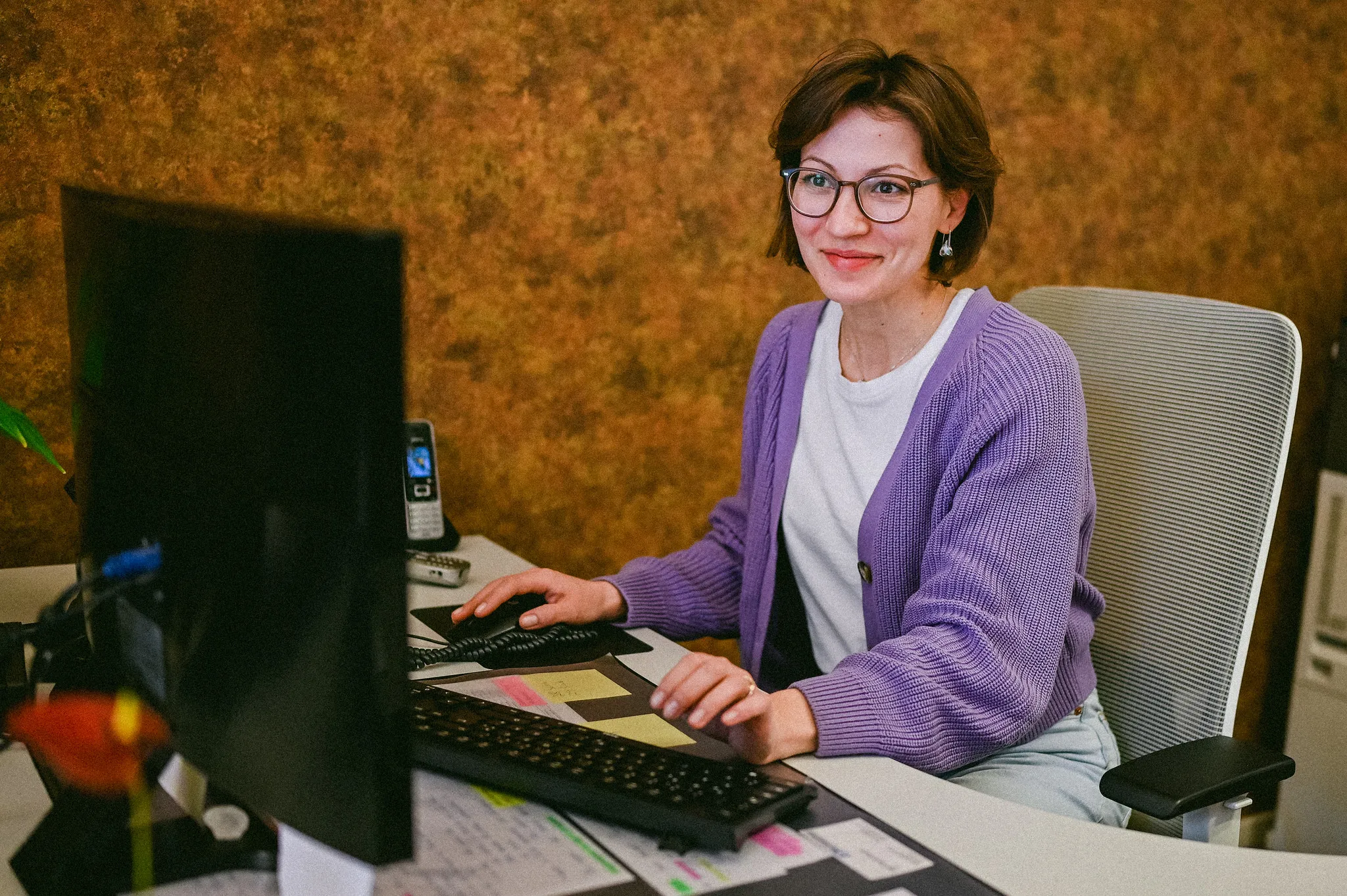 Frau mit Brille und lila Strickjacke am Schreibtisch, freundliches Business-Portrait