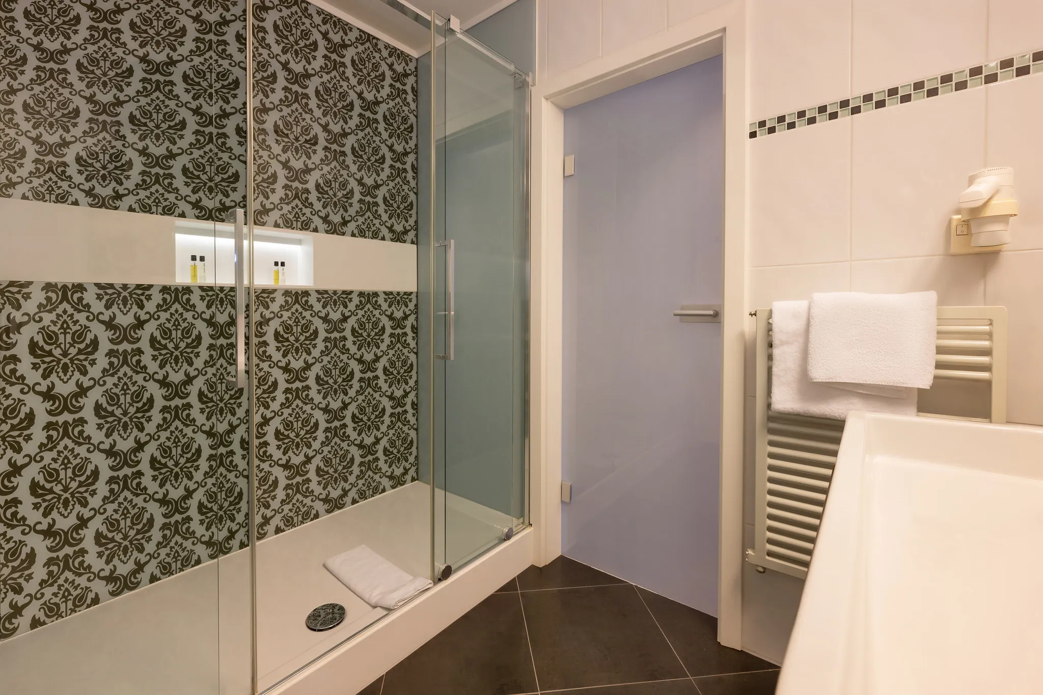 Hotelbadezimmer mit ornamentalen Motivfliesen, Glasdusche und Handtuchheizung