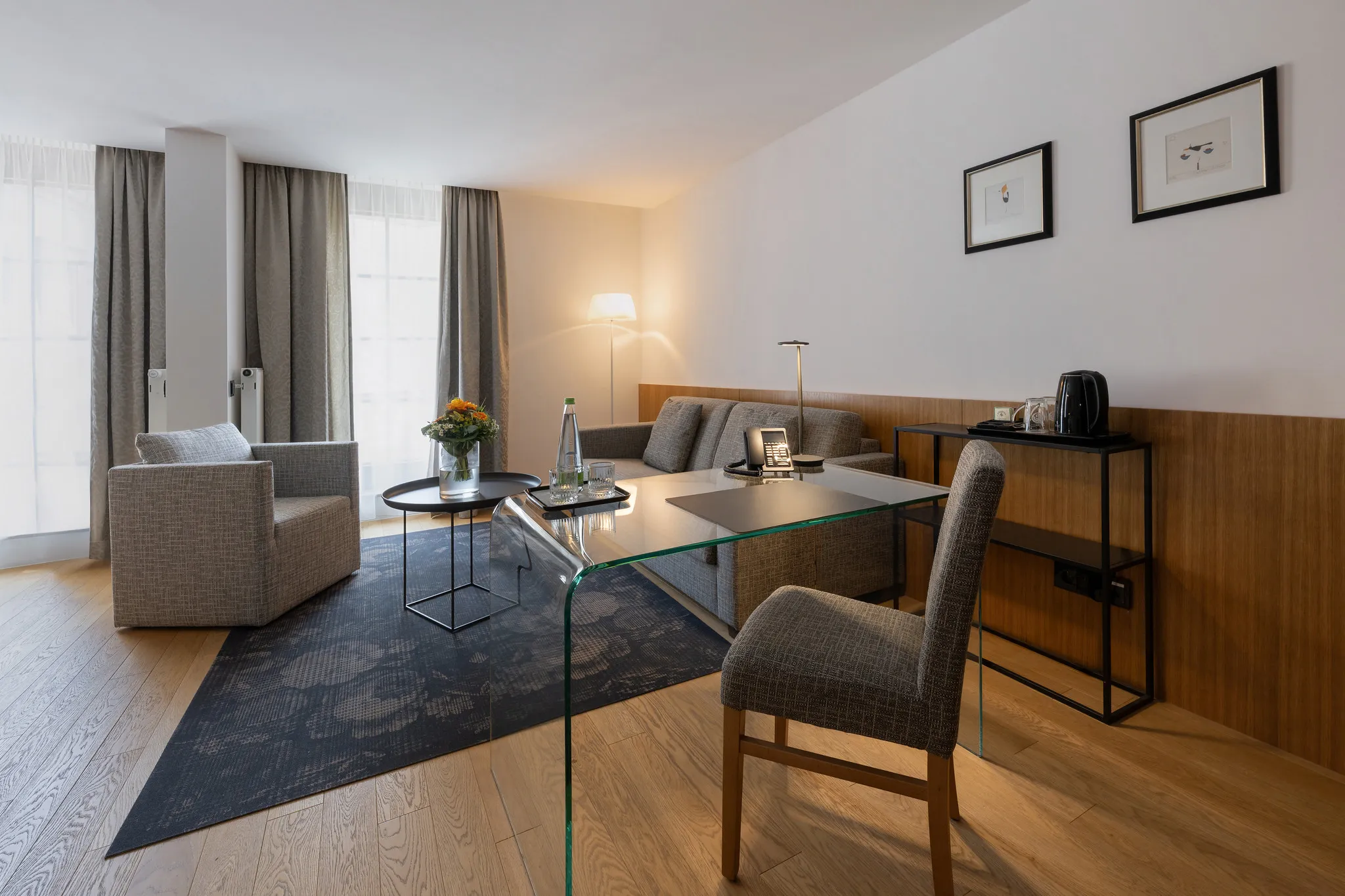 Helle Hotel-Junior-Suite mit Glasschreibtisch, grauem Sofa und großzügigem Sitzbereich