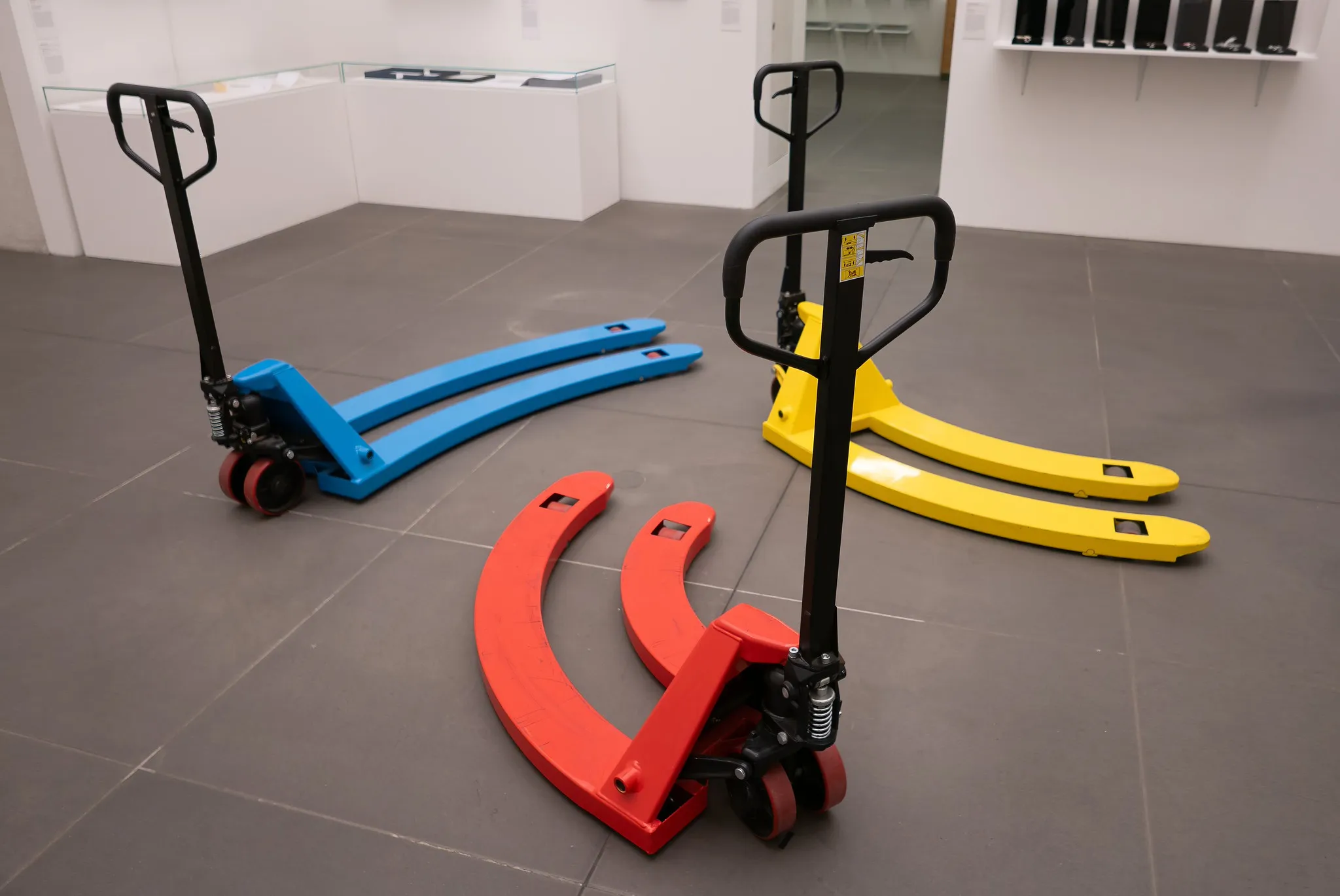 Drei farbige Hubwagen in Blau, Rot und Gelb im Galerieraum, lebendige Produktfotografie für Industrie-Equipment.