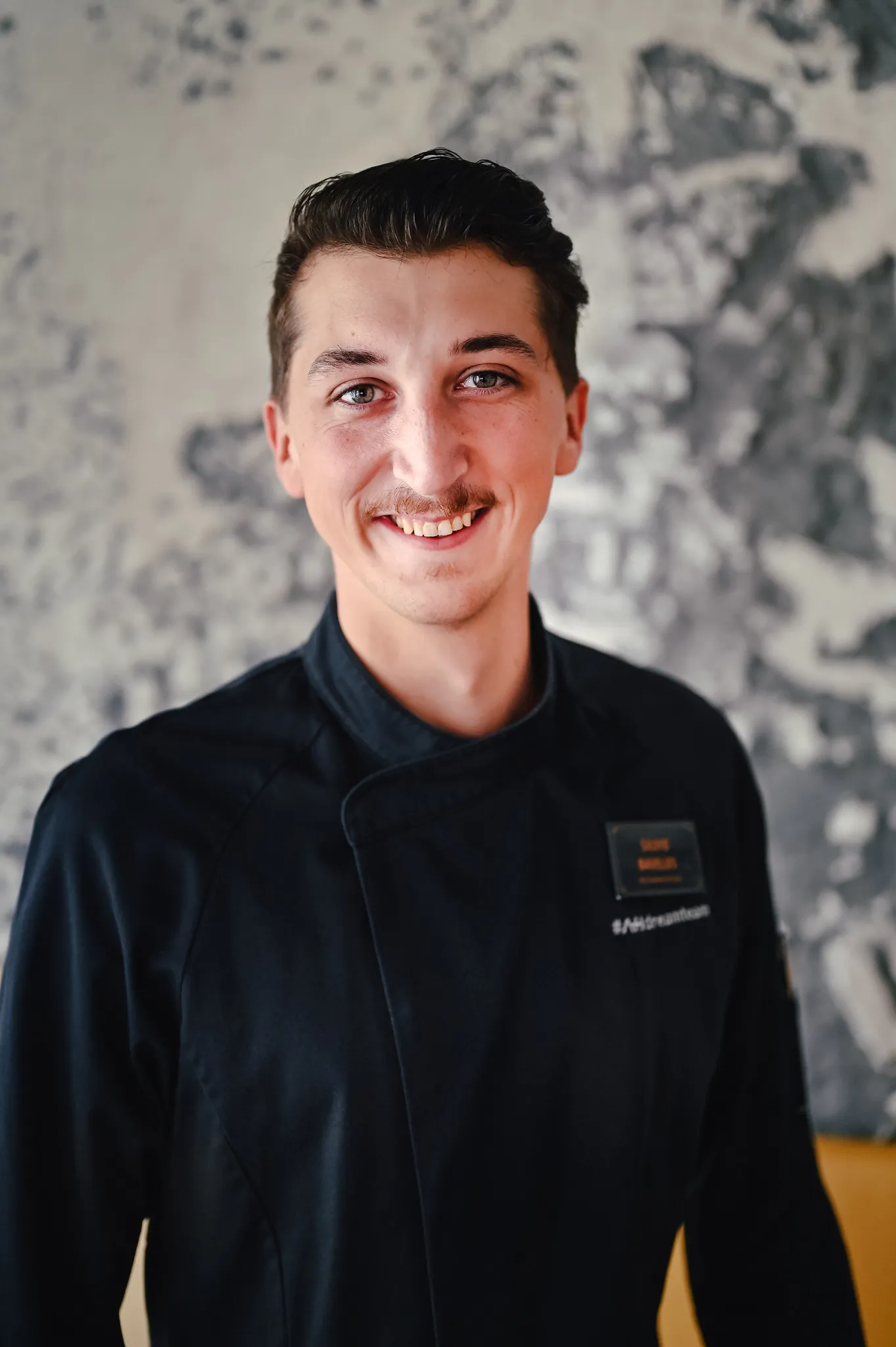 Junger Mann in schwarzer Kochjacke lächelt, freundliches Business-Portrait