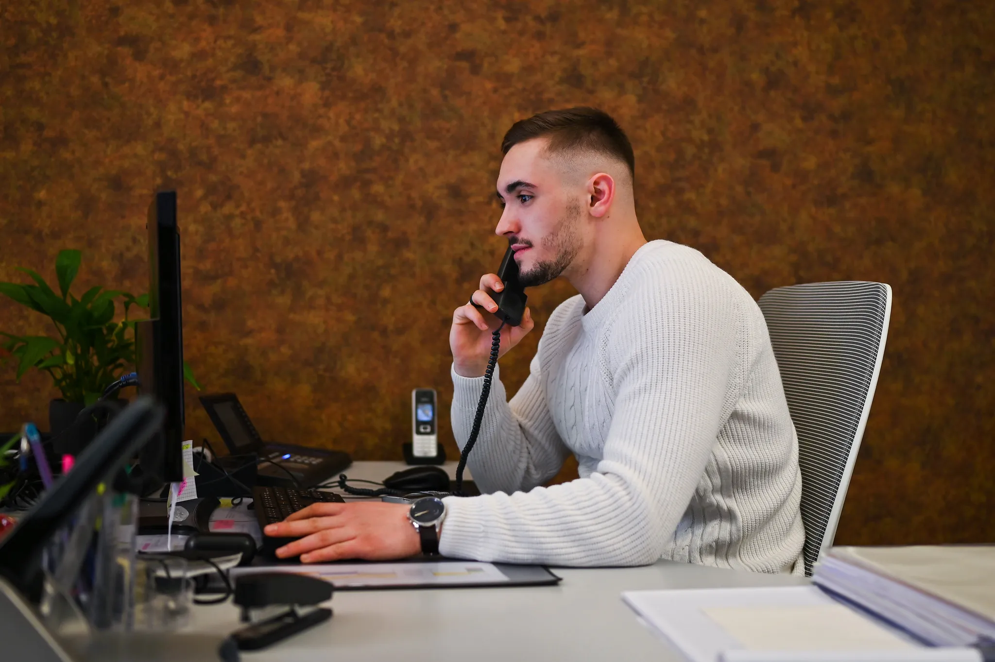 Junger Mann im weißen Pullover telefoniert am Schreibtisch, Business-Portrait im Arbeitsalltag