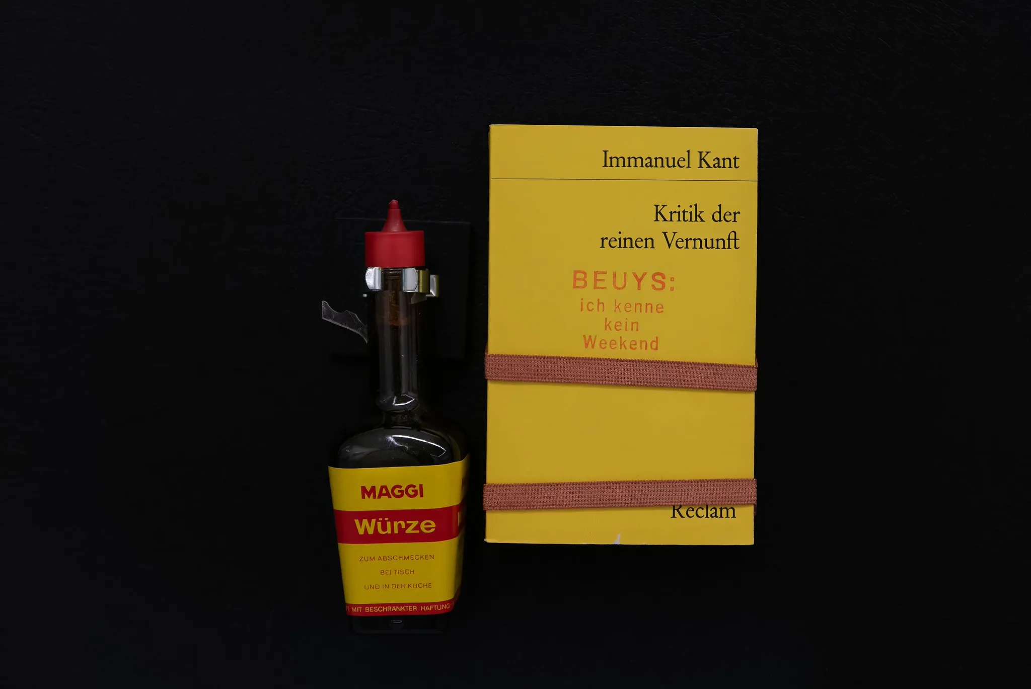 Maggi-Würze-Flasche neben gelbem Reclam-Buch auf schwarzem Hintergrund, kreative Produktinszenierung mit Kontrastwirkung.