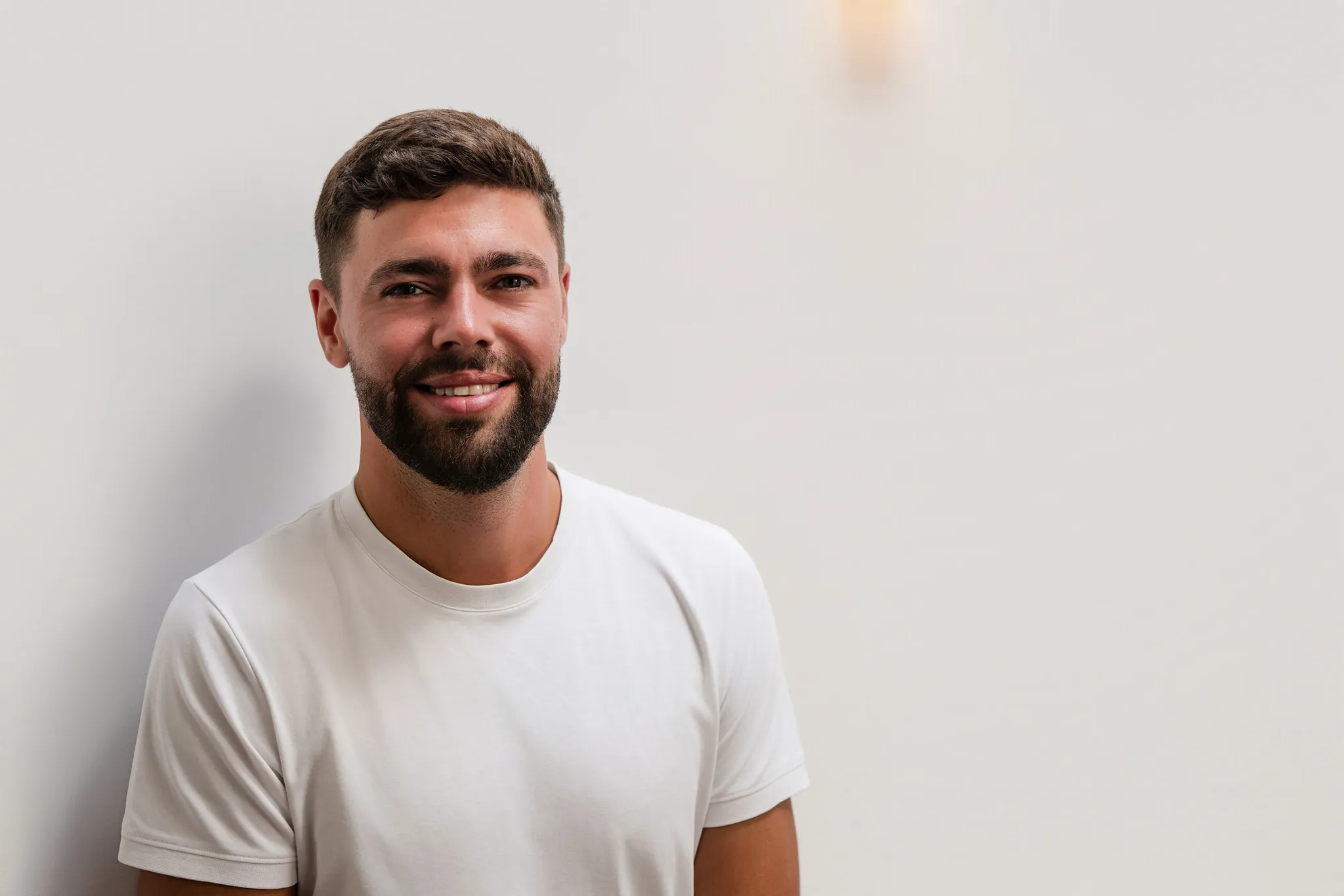 Mann mit weißem Shirt und Vollbart lächelt vor heller Wand, modernes Business-Portrait