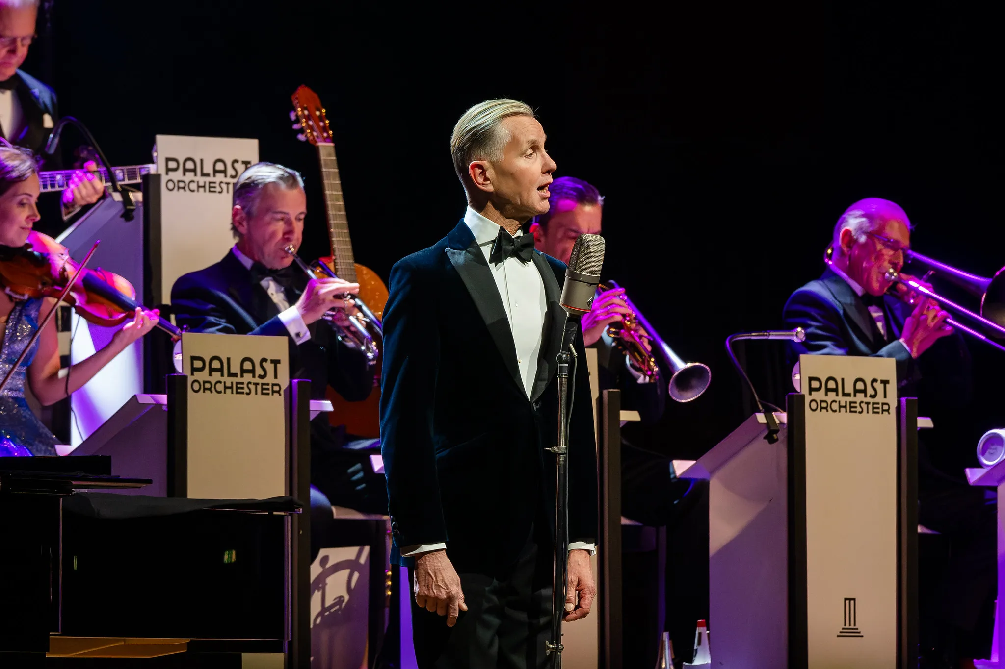 Solist des Palast Orchesters im Frack, Seitenansicht am Mikrofon, Geigen und Blechbläser im Hintergrund.