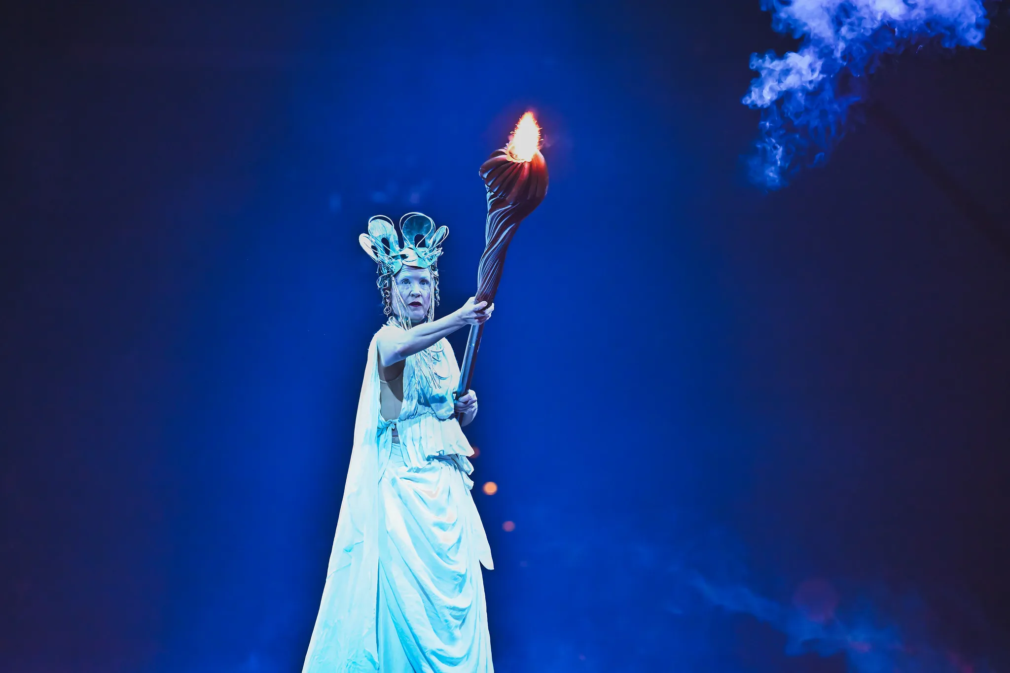 Theaterfigur in weißem Gewand und ornamentaler Krone hält eine brennende Fackel, blaues Bühnenlicht, Kulturfotografie.